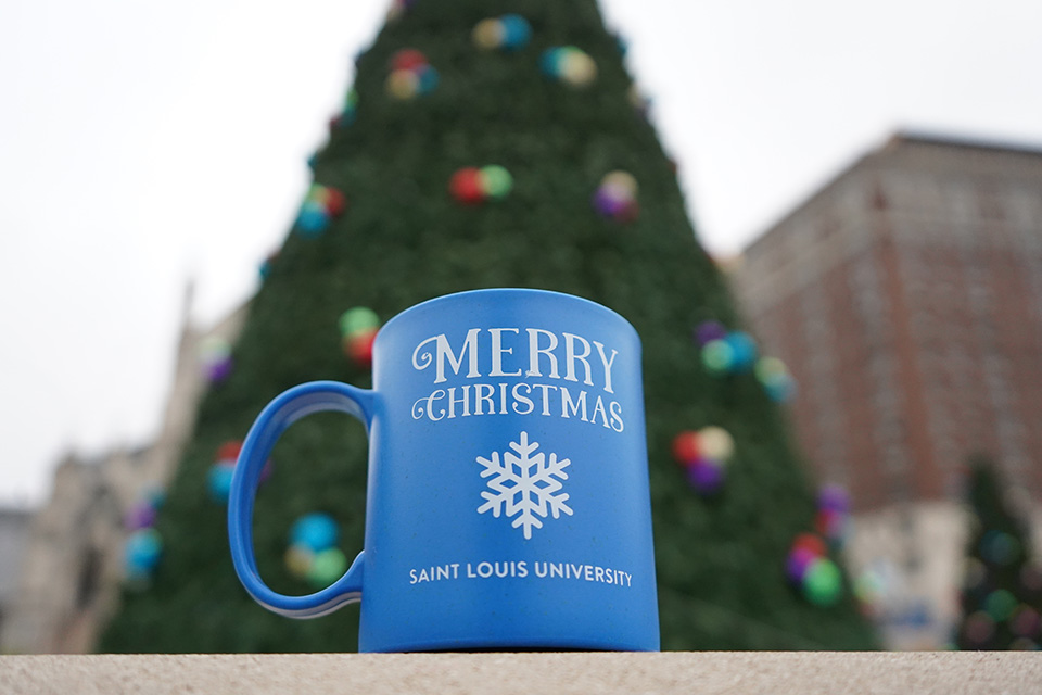 A mug says Merry Christmas Saint Louis University with an image of a snowflake on it as it rests in front of a large Christmas tree outside.