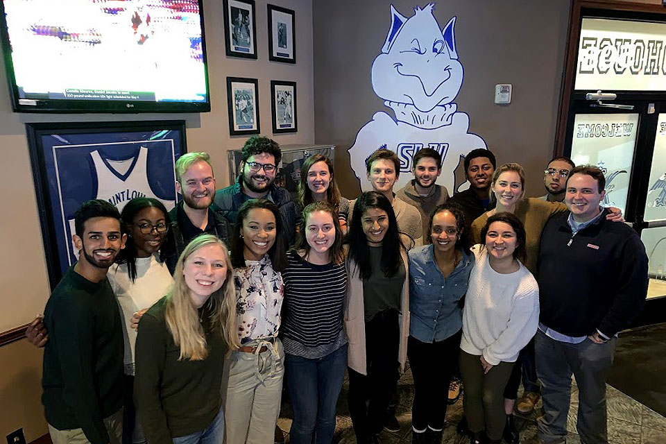 A group of students pose for a photo with a Billiken on the wall behind them.