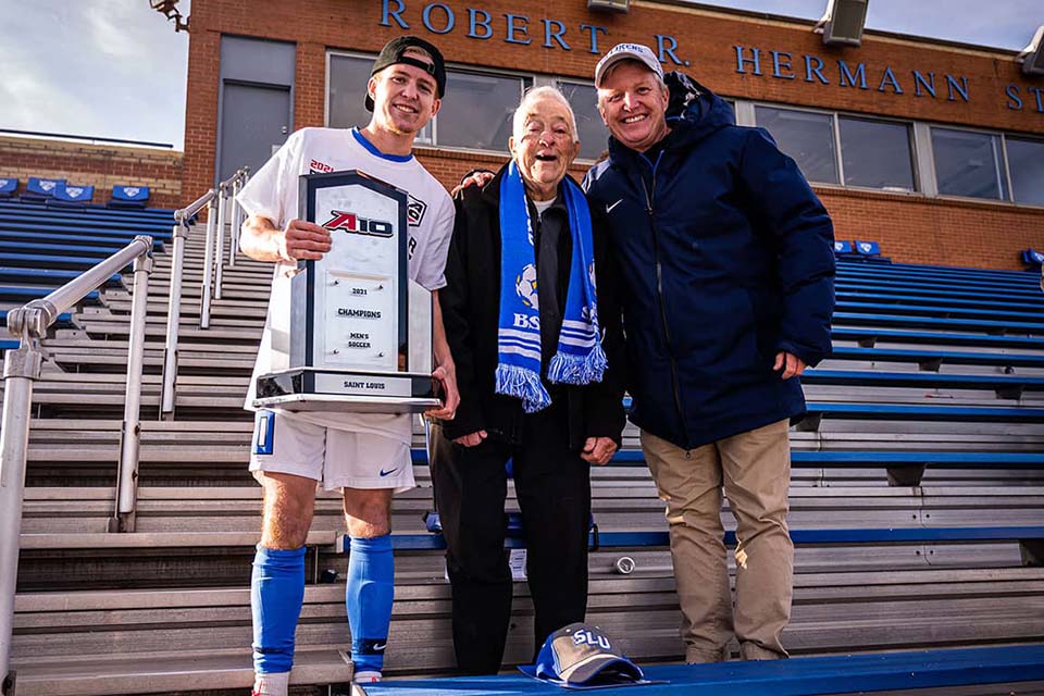 Three men pose for a photo together at the soccer stadium on campus.