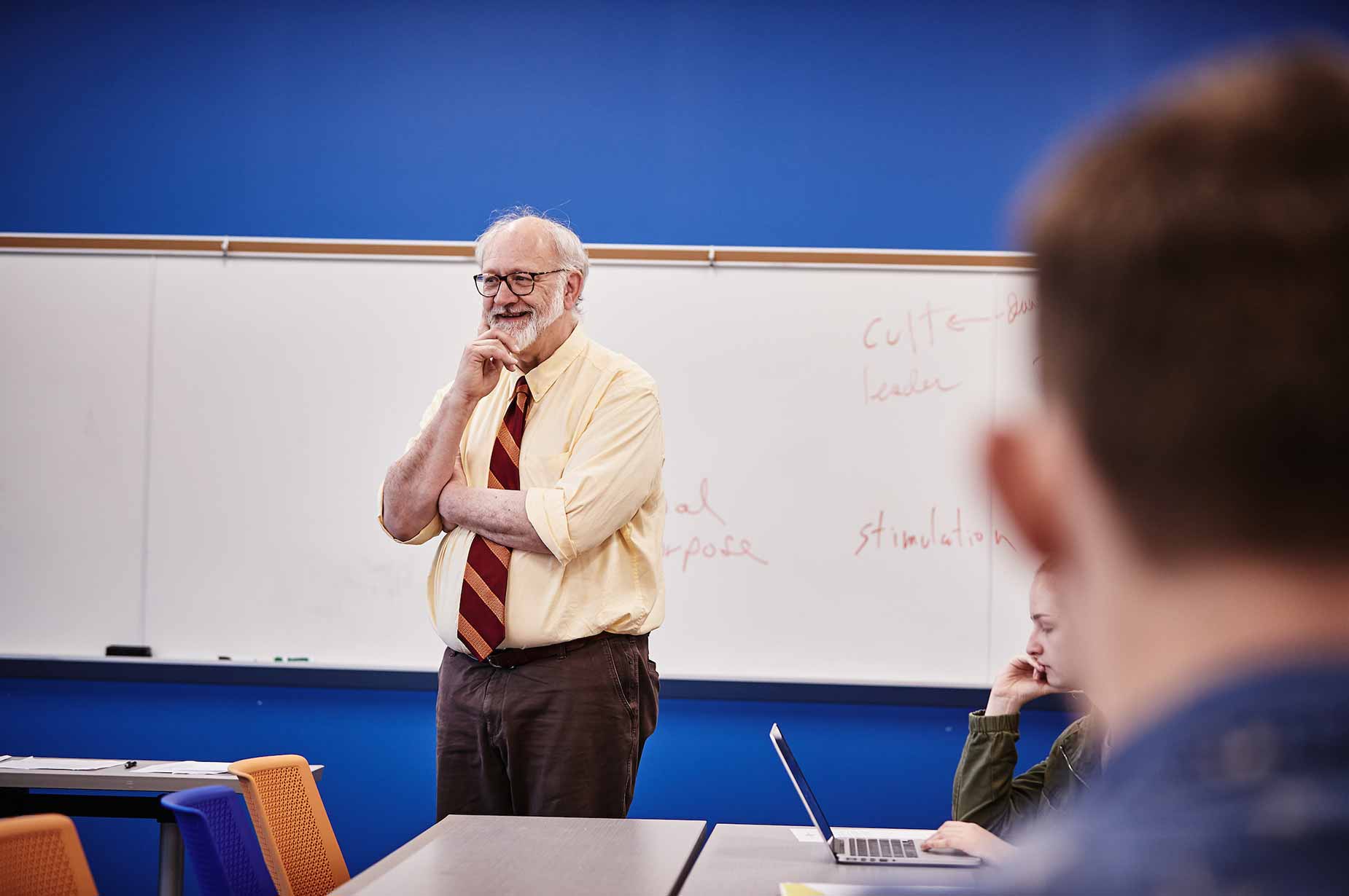 A male professor stands in front of his classroom. He is smiling and appears to be listening to someone speak.