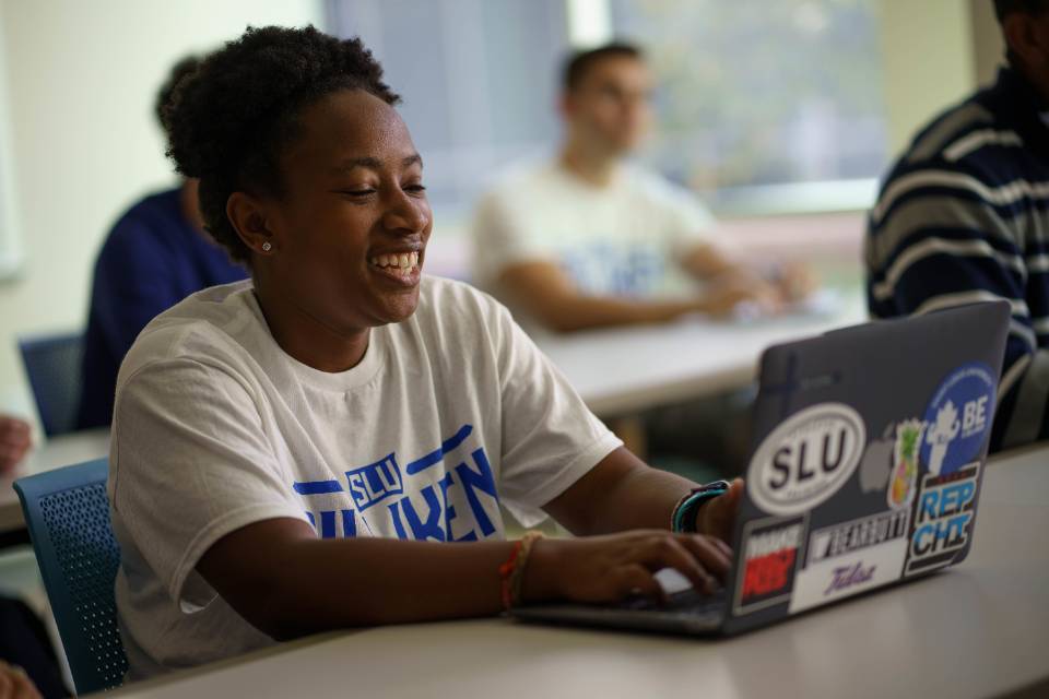 communication student in classroom A student wearing a Billiken t-shirt sits in a classroom, smiling while typing on a laptop.