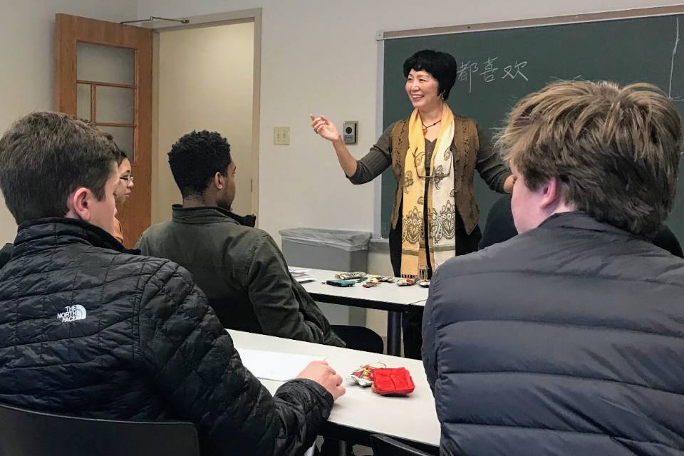 An instructor smiles while talking at the front of a classroom, as students are seen from behind. Chinese characters are visible on the board in the background.