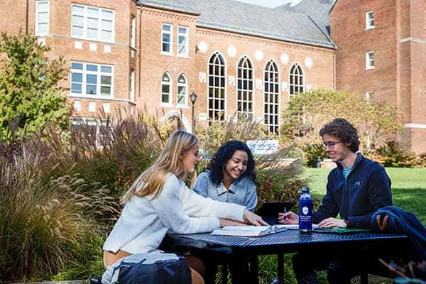 Three students work at a picnic table outside on SLU’s campus. Cook Hall can be seen behind them.