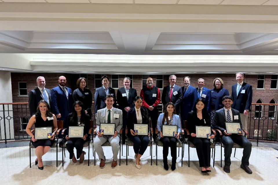 A group of students sit while holding awards plaques while professors and supporters stand behind them.