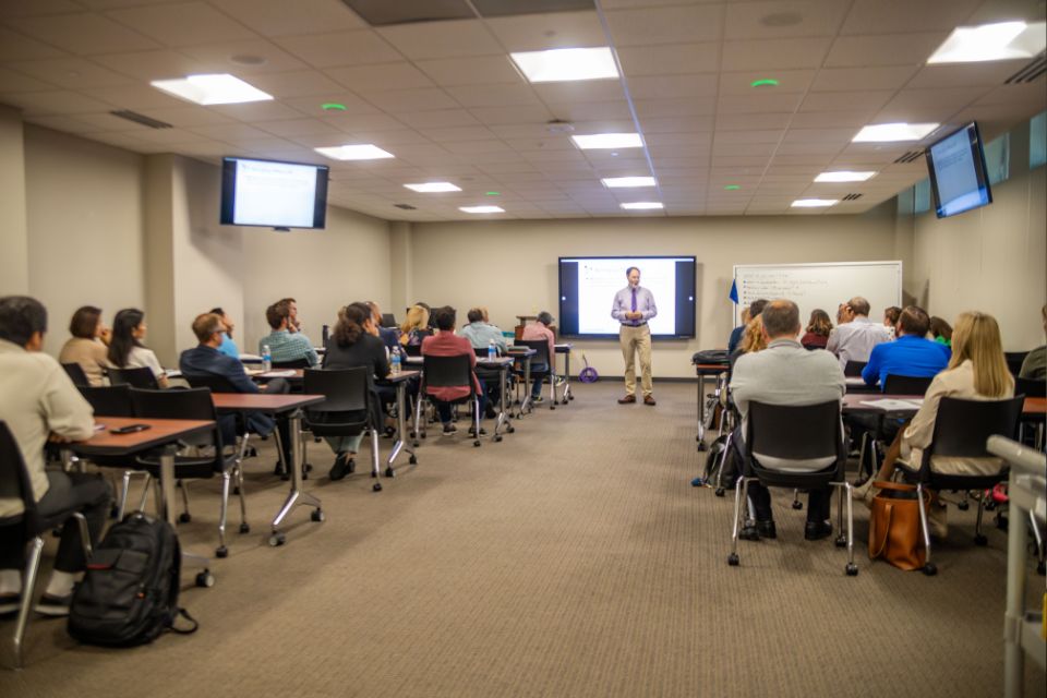 Participants seated at tables in a classroom setting, with a presenter standing in the front of the room delivering a presentation.