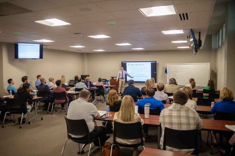 Participants seated at tables in a classroom setting, with a presenter standing in the front of the room delivering a presentation.