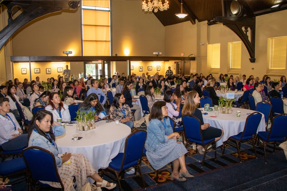 A crowded room of conference attendees are seen from the front while sitting at tables, watching a presentation