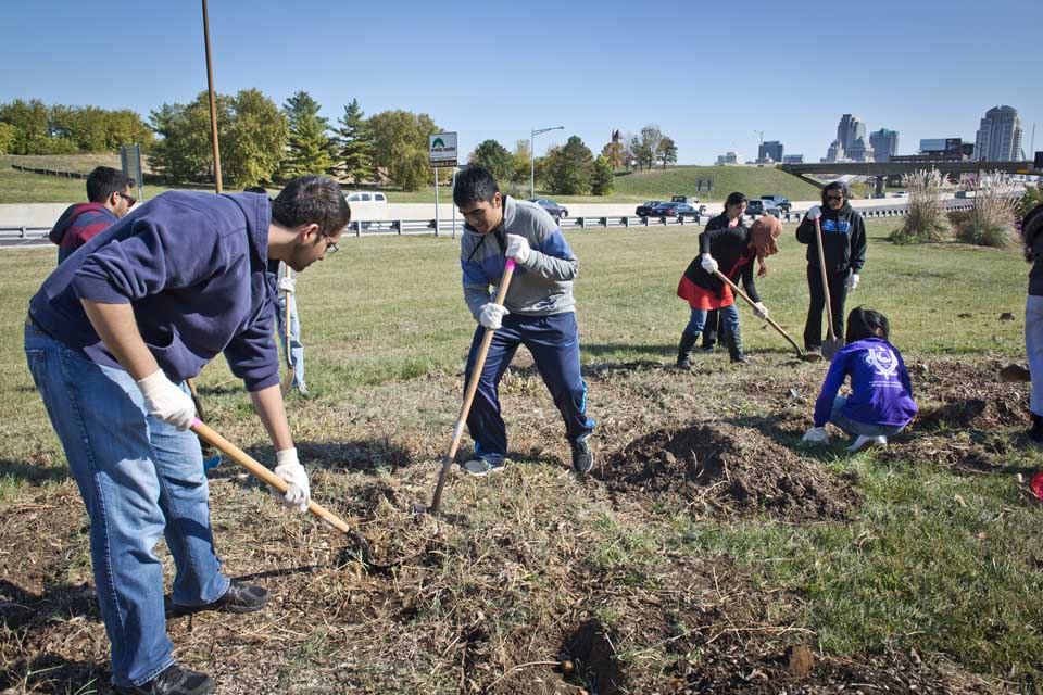 Students use shovels and other equipment in a grassy area next to a highway.