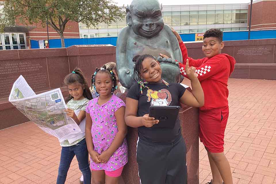 iScore students gathered around the Billiken statue, one holding a map, during a geocaching exercise