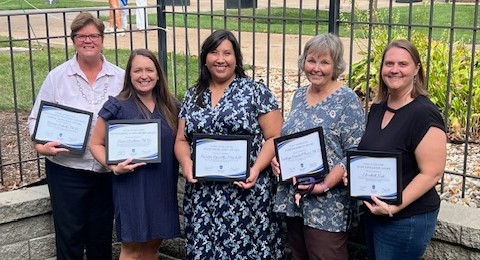 Five award winners pose side by side outdoors, holding their certificates.