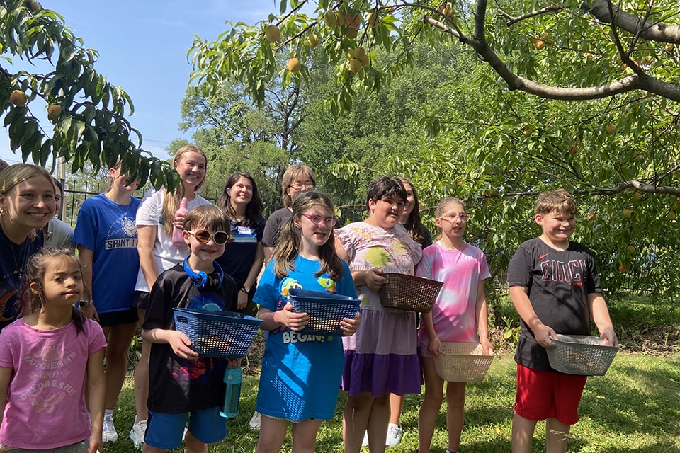 Students and camp staff stand under a peach tree holding baskets with peaches in them.
