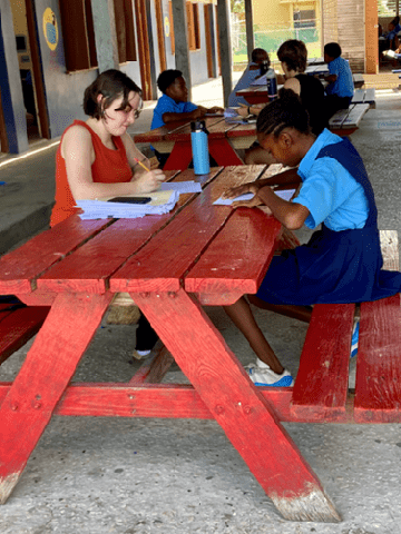 A SLU student sits at a picnic table with a younger student wearing a blue school uniform while looking at school work