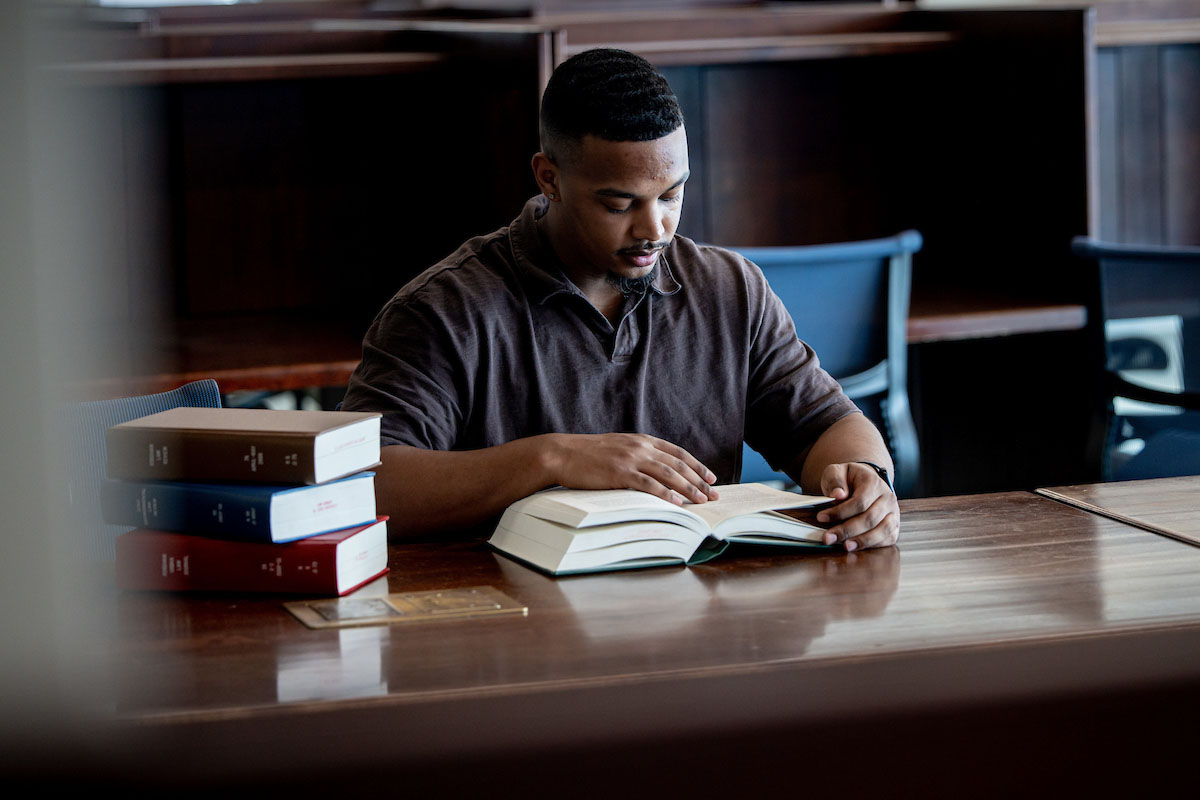 A student reads a book at a wood table in the library with a stack of books on the table next to him.