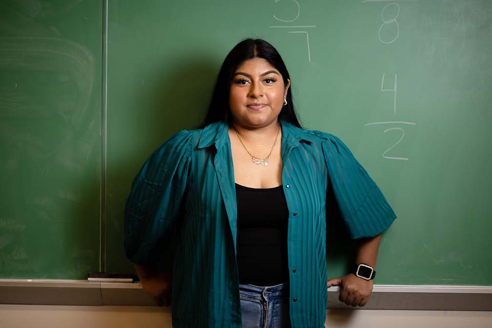 A student poses in front of a chalkboard with arithmetic problems written on it.