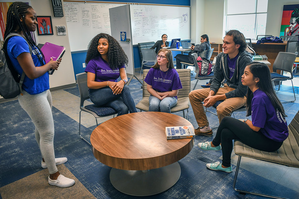 Four students, each wearing t-shirts that read "live. learn. connect." sit around a round coffee table while looking up at another student wearing a SLU Billikens T-shirt.