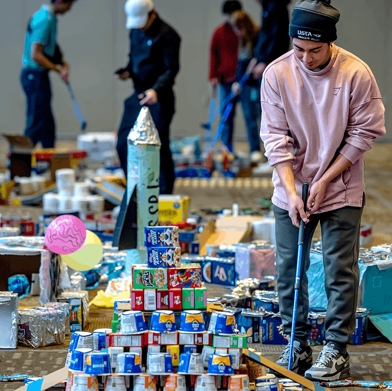 A student gets ready to putt a golf ball next to a pyramid made out of food packages.