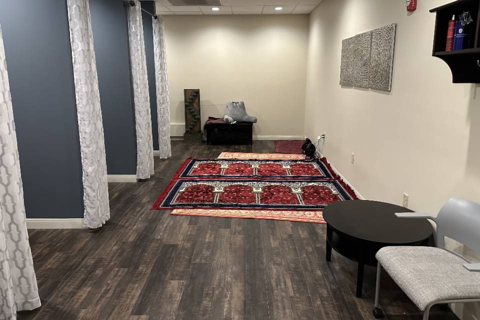 A long narrow room with alcoves on each side with curtains. There are prayer rugs lined up on the floor, as well as some chairs and cushions.