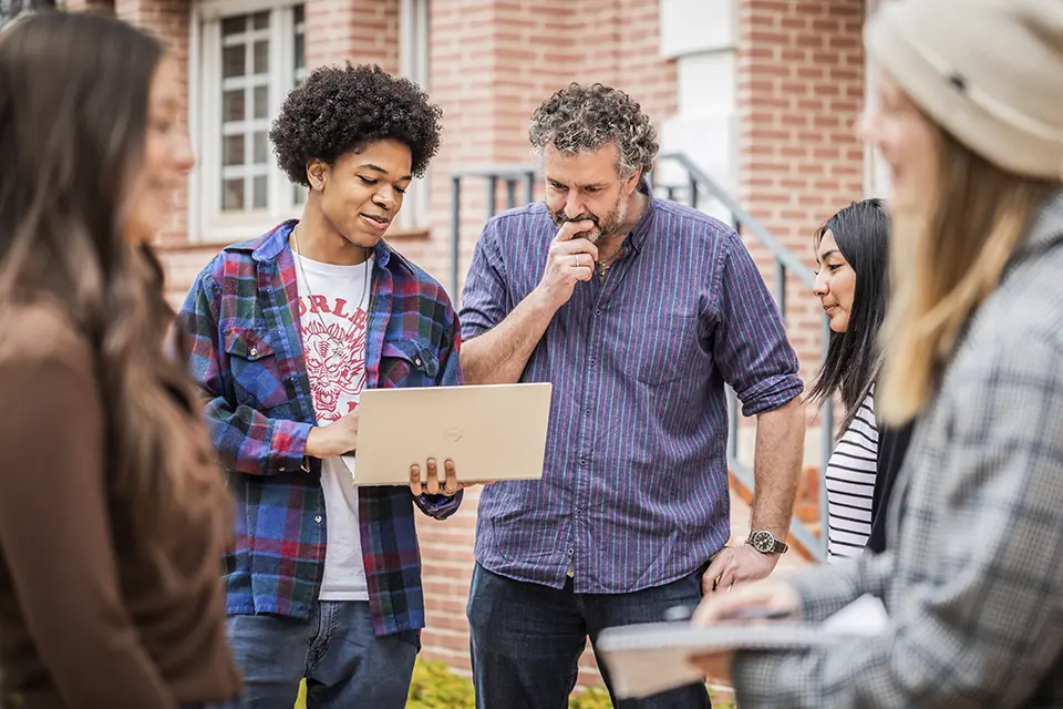 Professor with students looking at a laptop while standing outside.