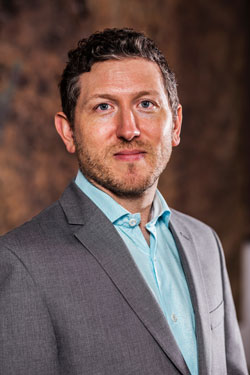 MOCRA Director David Brinker, wearing a light blue dress shirt and grey sportcoat, poses in front of an abstract brown background