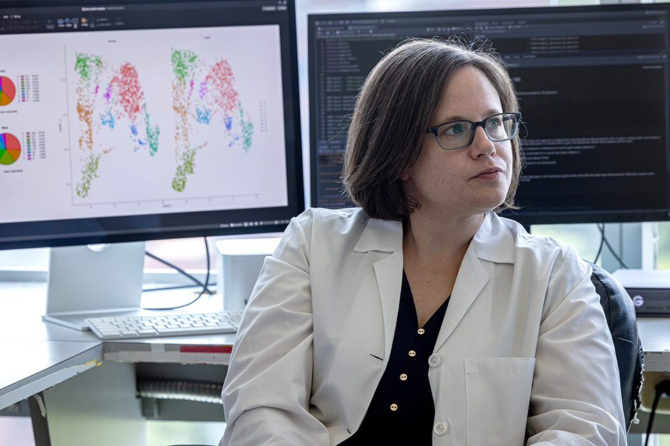 A researcher in a white coat sits and looks off to the side with a computer screen displaying data behind her.