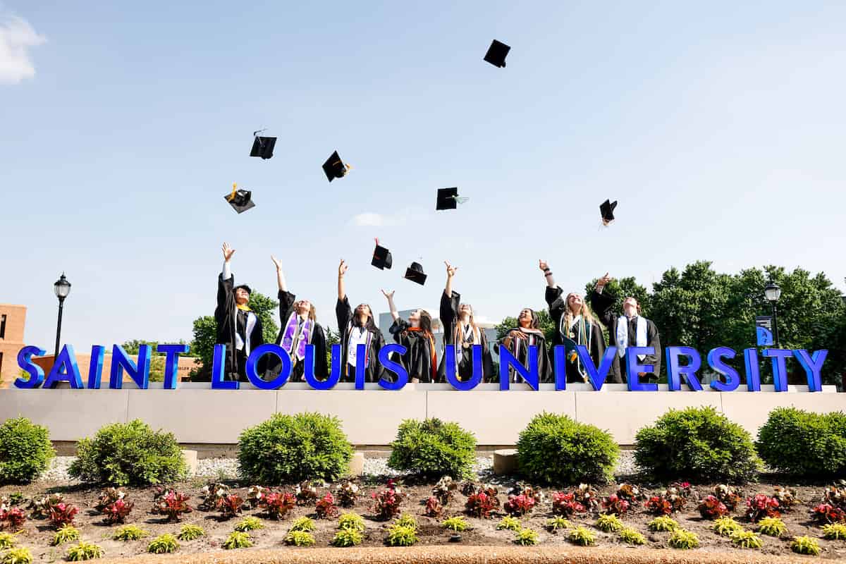 SLU graduates throw their caps behind the Saint Louis University sign at the corner of Lindell Avenue and Grand Boulevard.