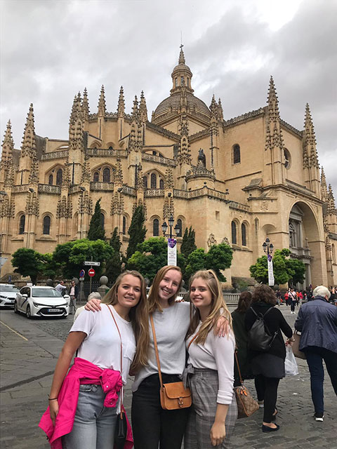 Three students stand with their arms around each other with a castle-type building in the background.