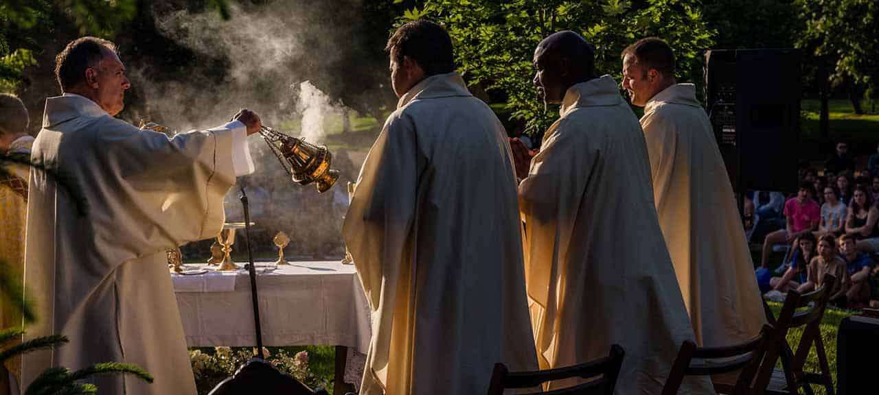 A man wearing white vestments swings a censer over the alter while celebrating Mass. Three other men in vestments look on.