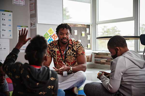 A male student sits on the floor of a classroom with a group of children. A young girl is raising her hand to speak.