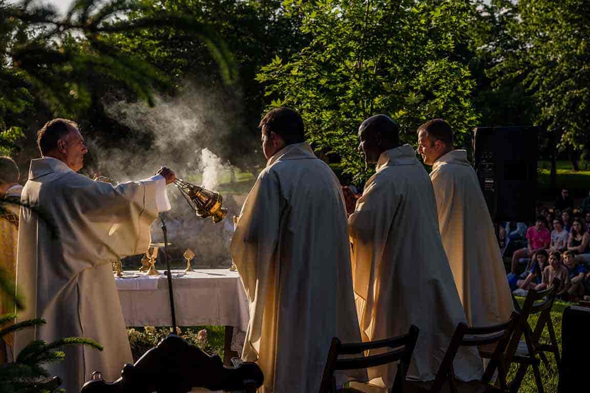 Celebrants in liturgical vestments stand alongside an outside alter while another celebrant swings incense over the alter.