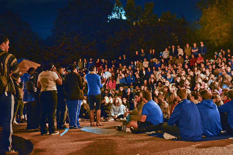 Students fill the steps of Lipic clock tower and listen as a speaker talks to the group. It's night time and street lights light the scene.
