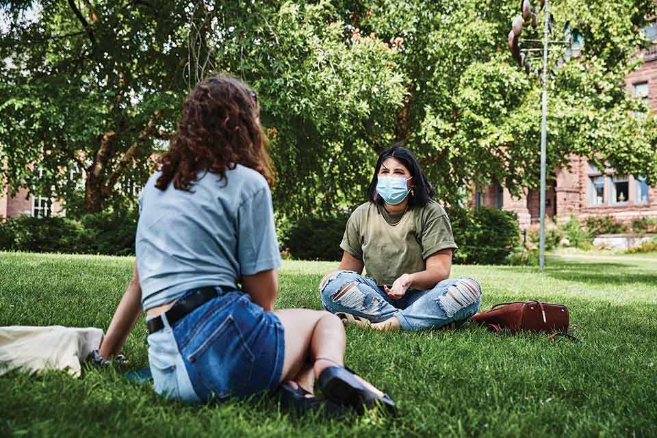 Two female students sit and talk six feet apart on SLU's quad. They are both wearing medical masks.