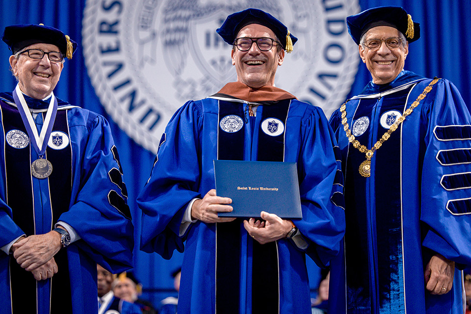 Actor Jon Hamm smiles as he holds a SLU diploma during the 2024 commencement ceremony. He is flanked by Joe Conran (left) and Fred Pestello (right). All men wear royal blue academic regalia.