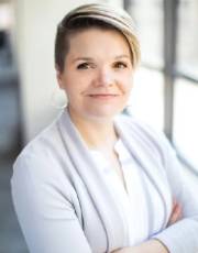 Katie Heiden Rootes headshot next to window with arms folded.